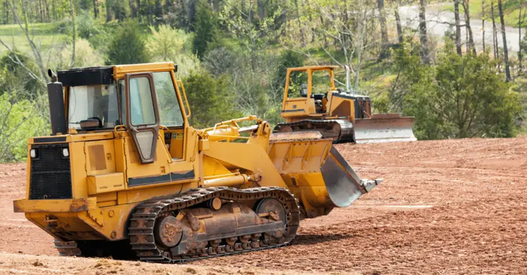 Two yellow bulldozers on a cleared patch of bare red dirt with trees behind