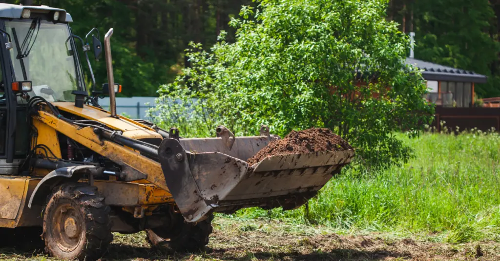 Backhoe loader bucket carrying soil across a cleared grassy lot