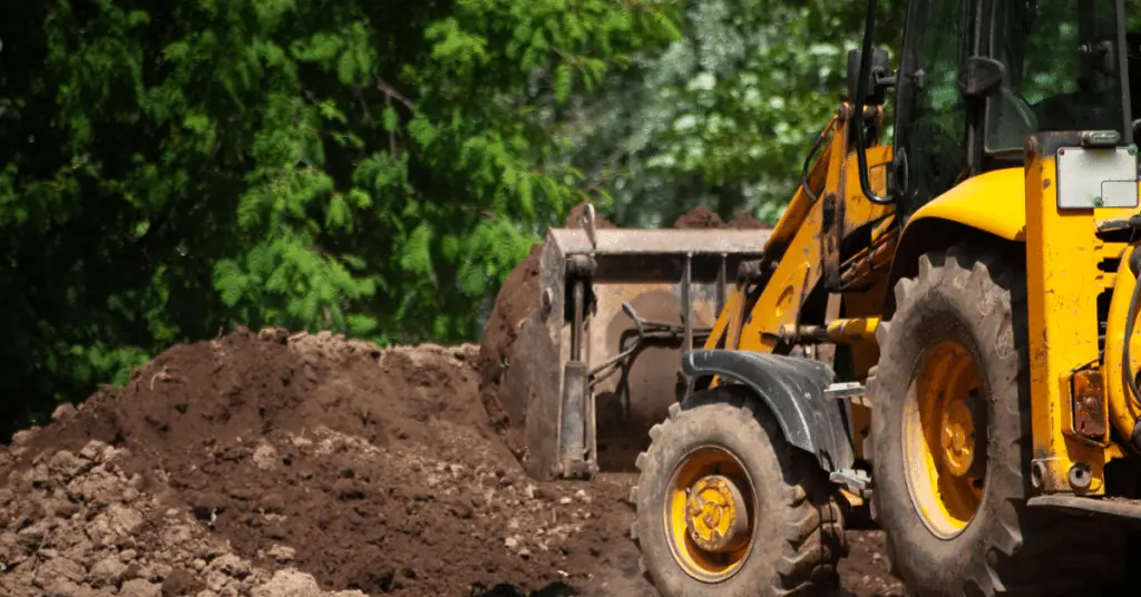 Yellow backhoe loader beside a large pile of dirt in front of green trees