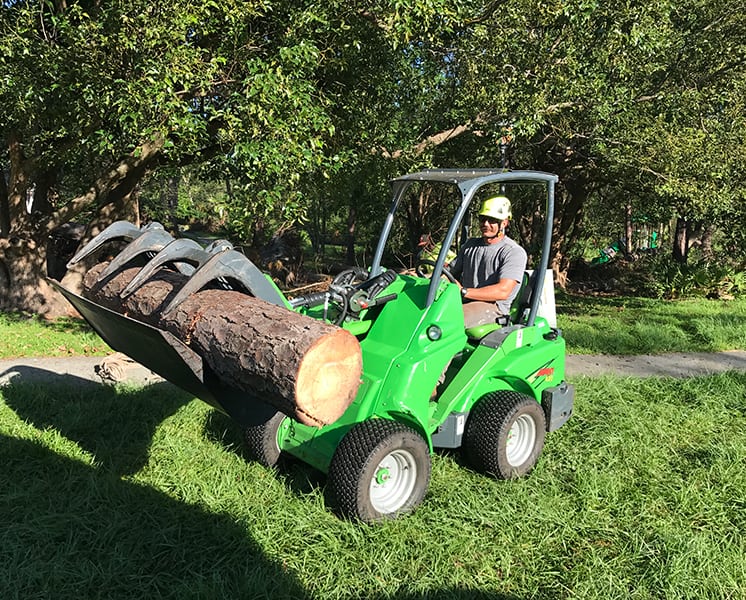 Worker driving a green mini loader with a grapple that carries a large log across a lawn